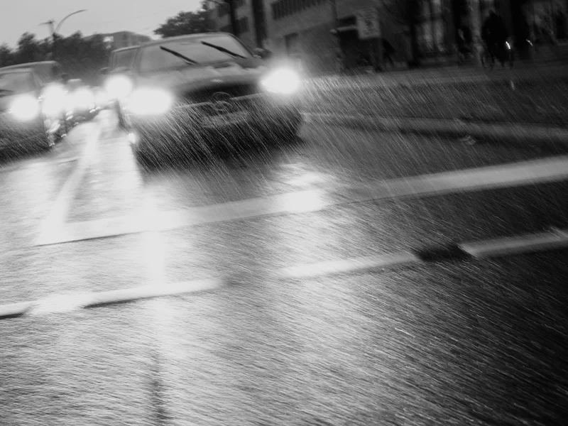Black and white photo of cars on a rainy street with blurred headlights in front of a tall building.