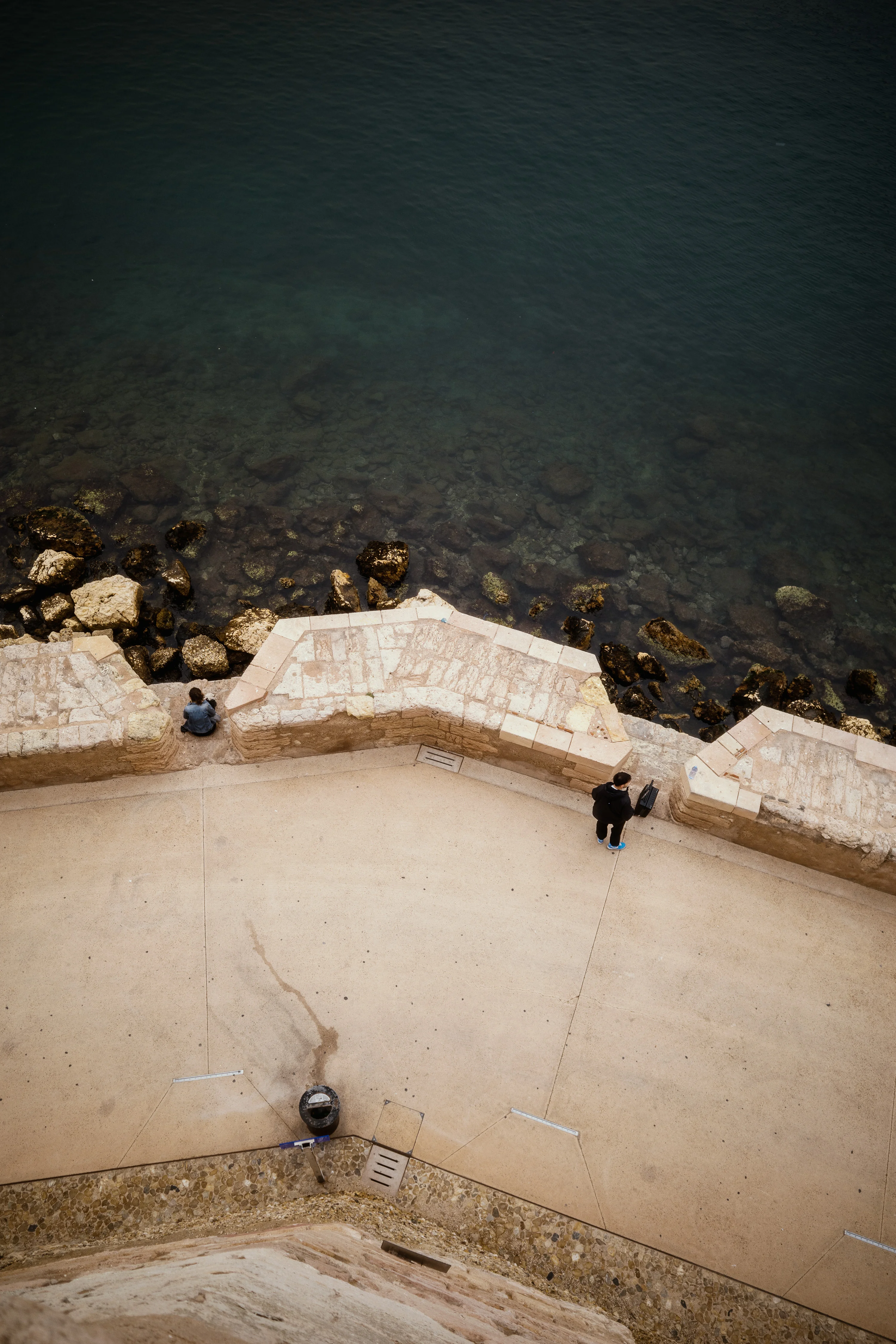 Two people by a stone wall overlooking a rocky shoreline with a wide expanse of water.