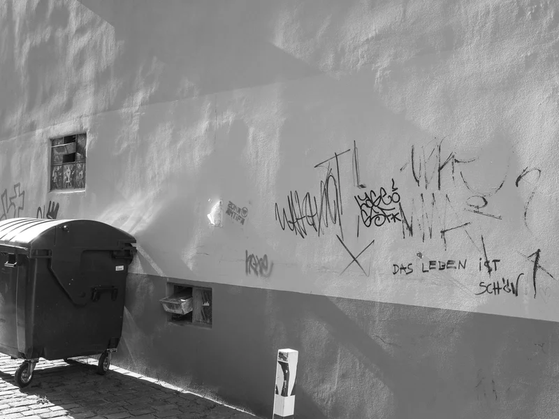 A wall with various graffiti tags and a garbage bin on a cobblestone street.
