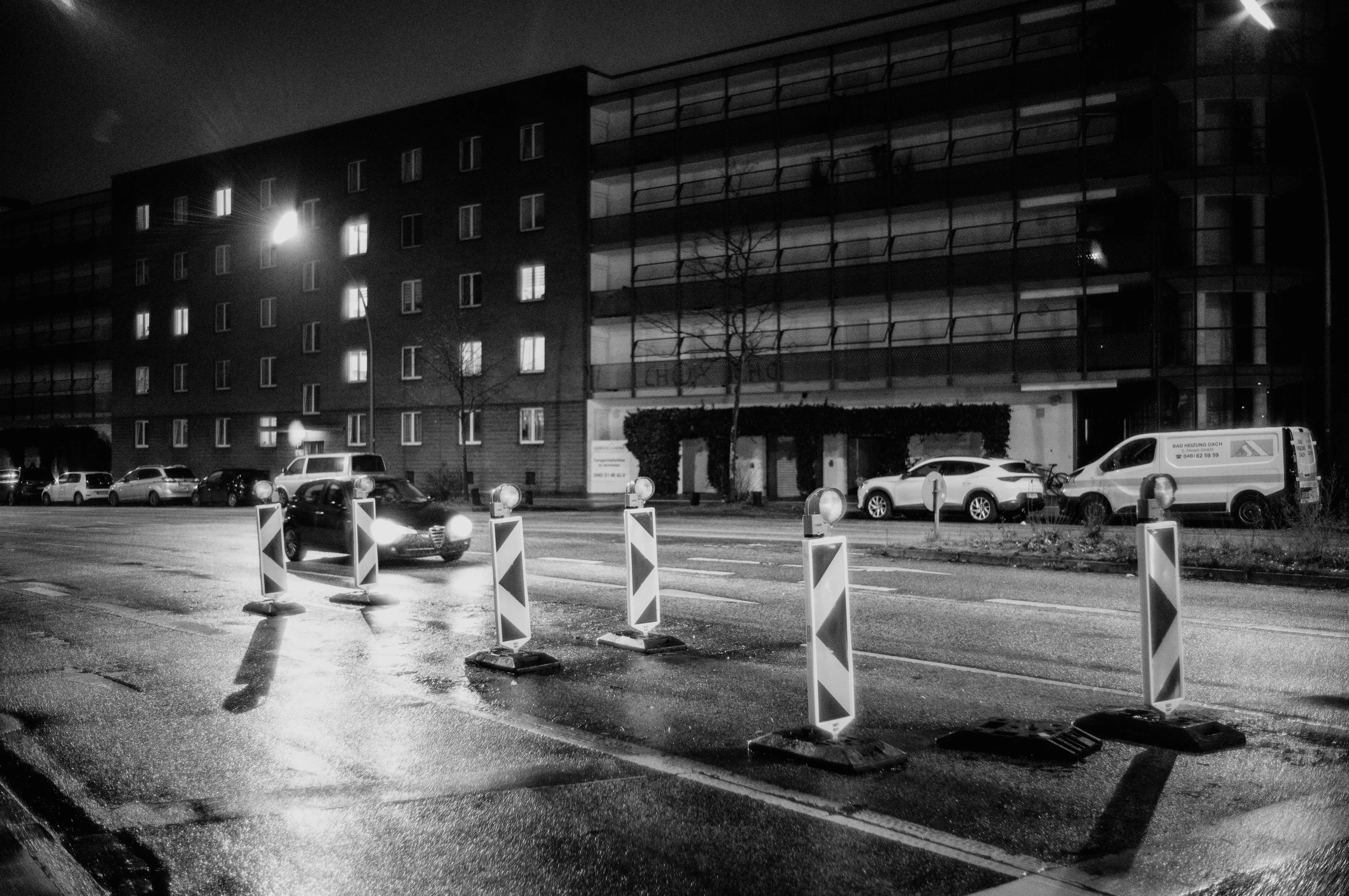 A nighttime street scene featuring a car driving past road barriers in front of a dimly lit building.