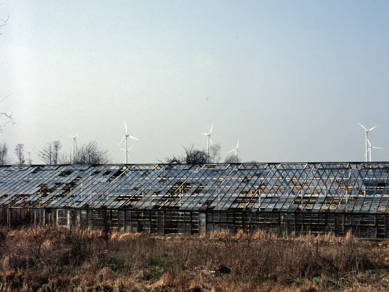 A dilapidated greenhouse with wind turbines in the background under a clear sky.