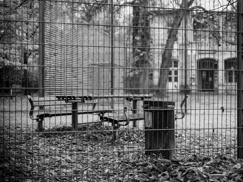 Black and white photo of an outdoor area with benches and a trash can behind a metal fence.