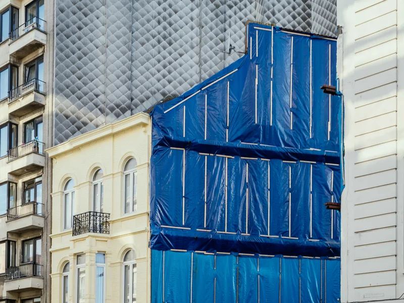 A building facade under construction with blue tarpaulin covering next to a yellow and a white building.