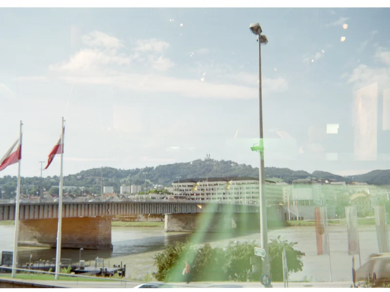 Cityscape with a bridge over a river, flags, and a hillside in the background.