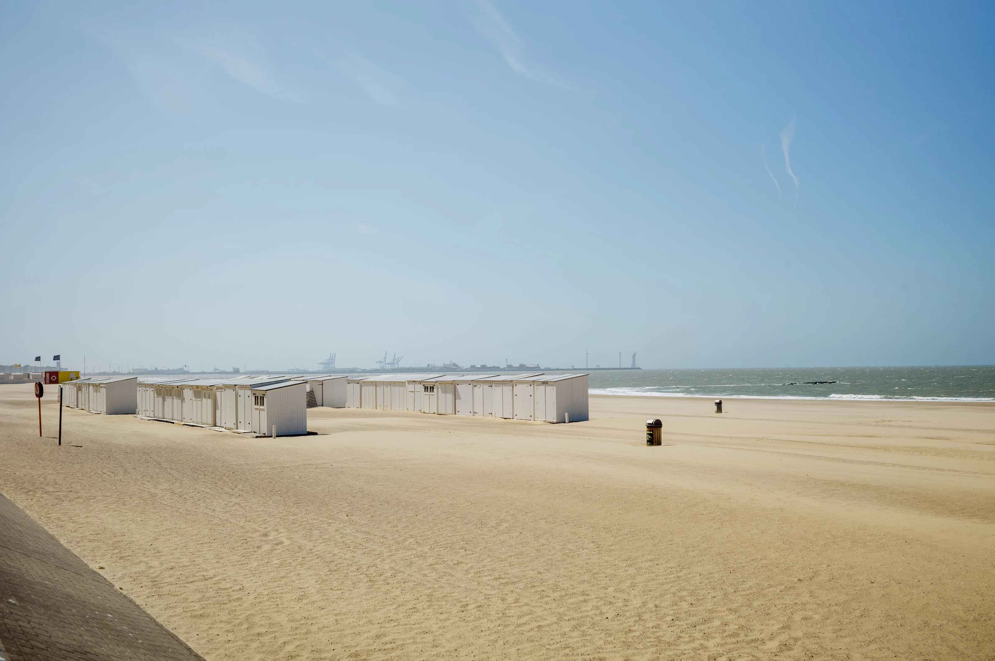 Sandy beach with row of white cabins near the shoreline under a clear blue sky.
