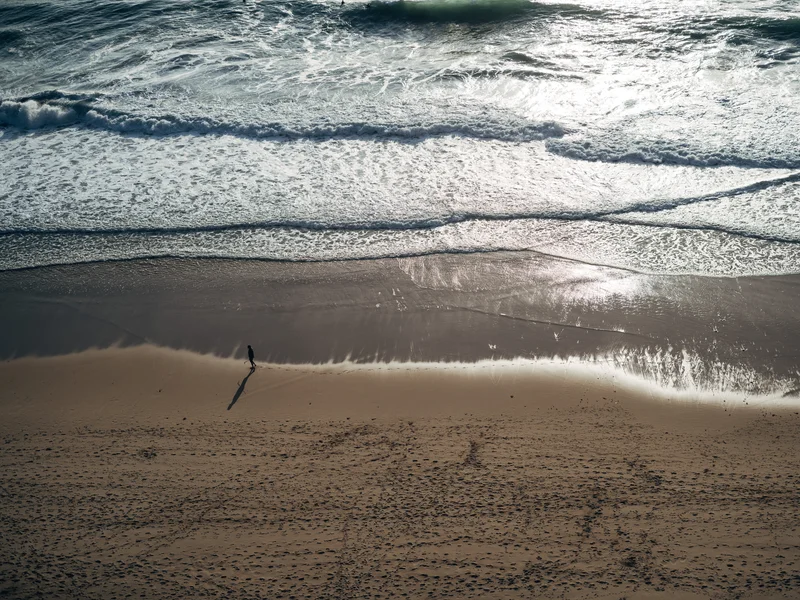 A lone person walks along a sandy beach with ocean waves rolling in.