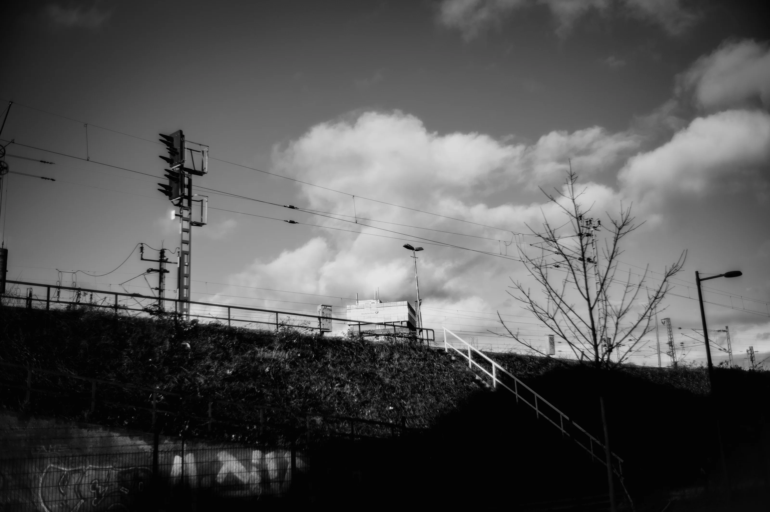 Black and white photo of a railway signal and power lines with stairs leading up a grassy slope against a cloudy sky.