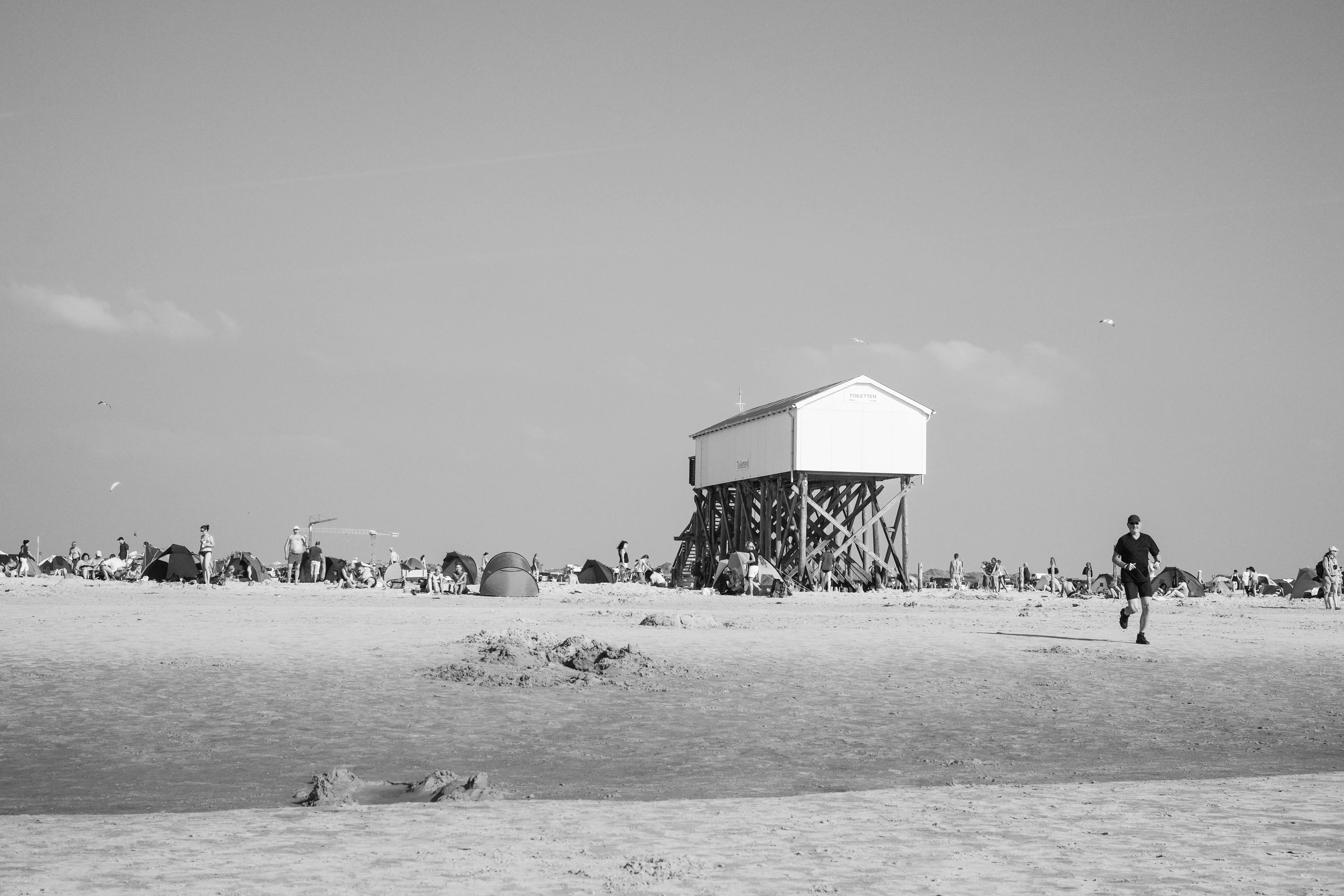A black and white photo of a beach scene with a raised building and people enjoying the shore.