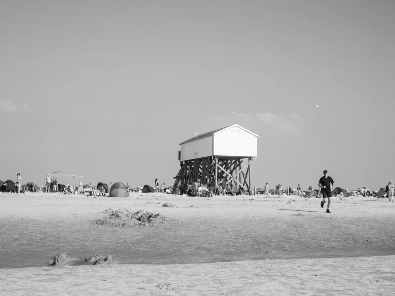 A black and white photo of a beach scene with a raised building and people enjoying the shore.