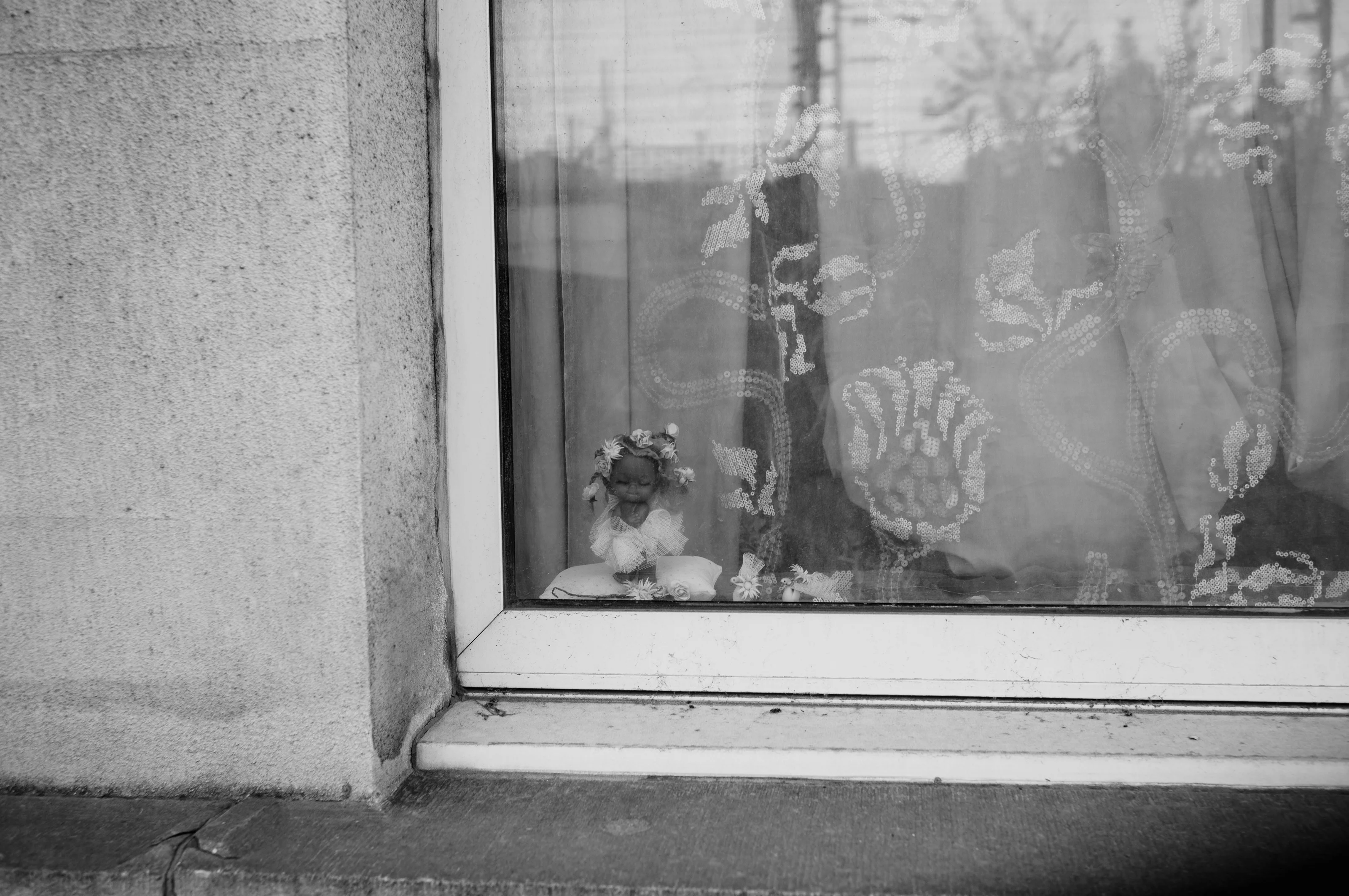 A small doll with a floral crown is visible through a lace curtain in a window.