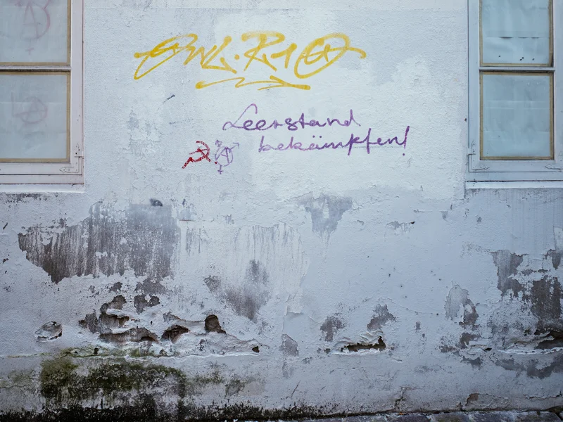 Graffiti on a weathered wall between two windows with cracked paint.