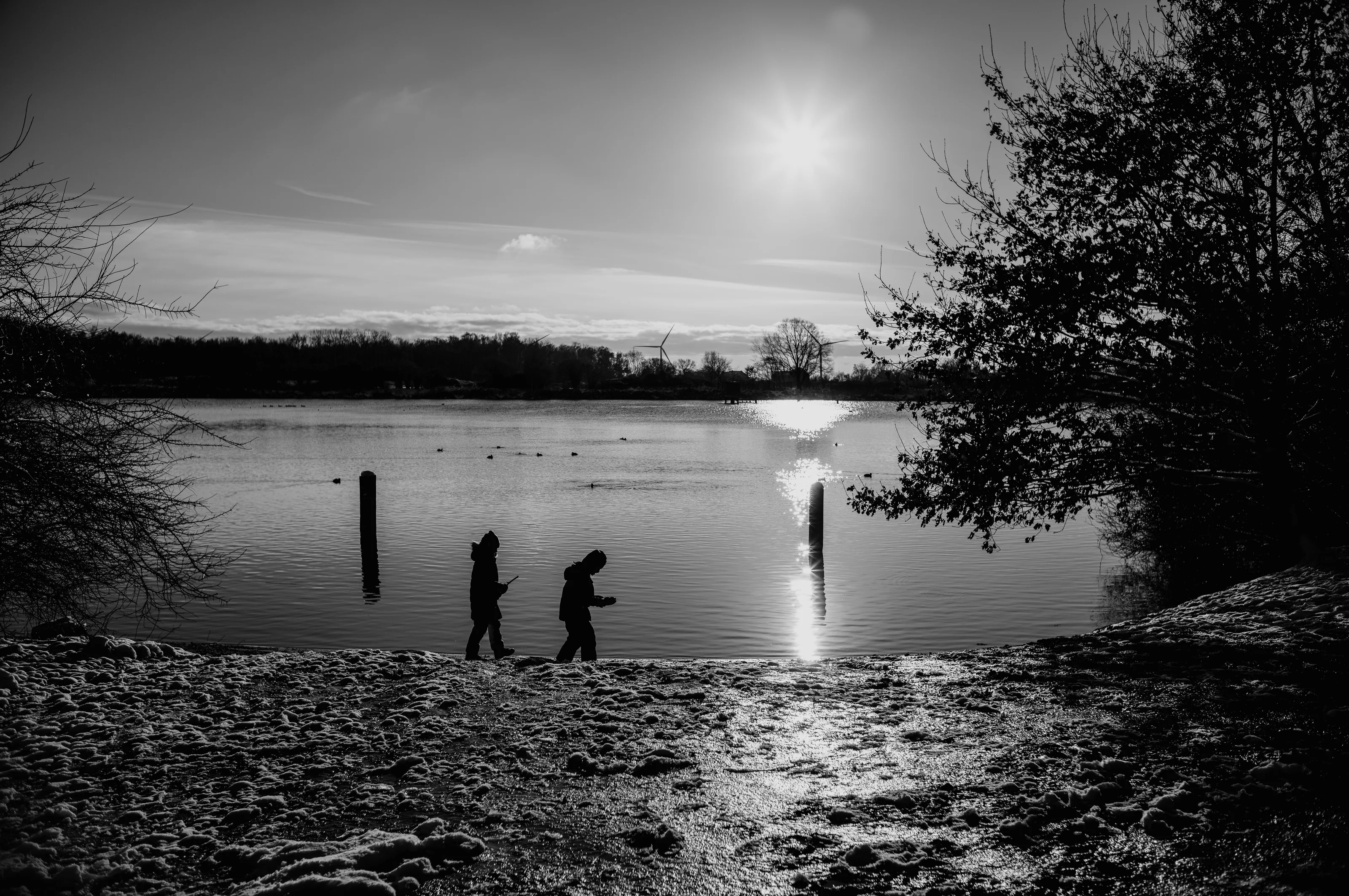 Two people walking near a lake with a bright sun reflecting on the water.
