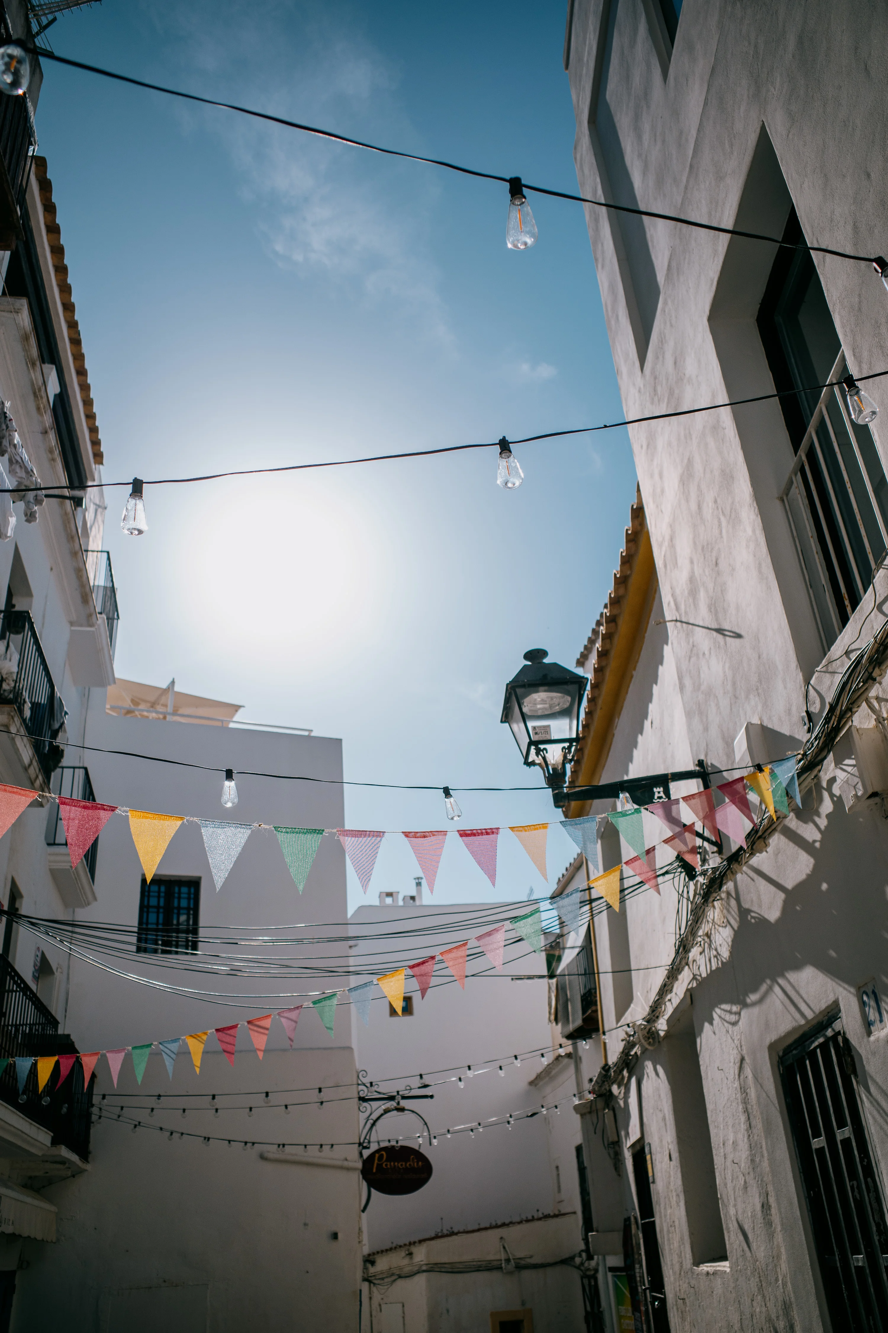 Narrow alleyway with colorful bunting and light bulbs strung between buildings under a clear blue sky.