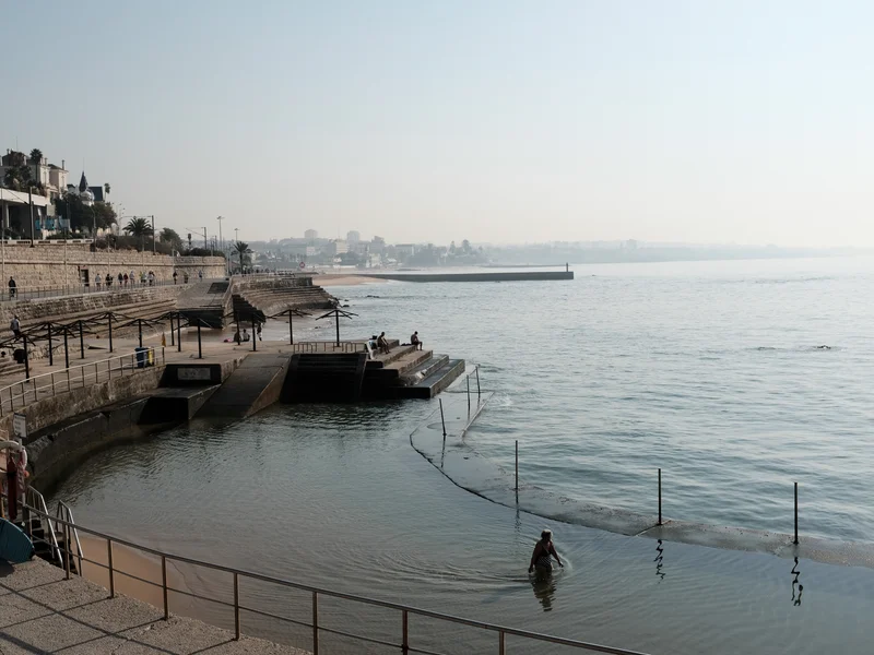 Seaside promenade with people walking and a person wading in the water near steps and railings.