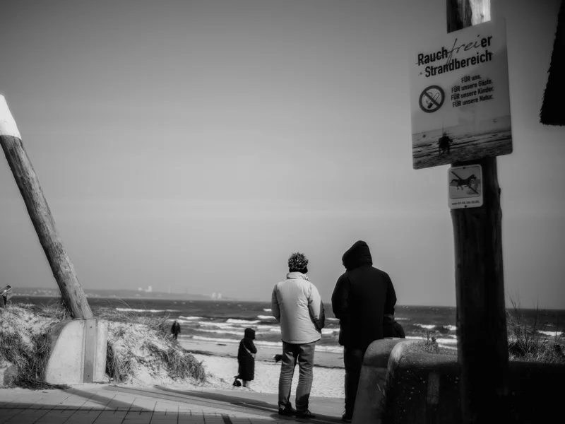 Two people in jackets stand on a beach walkway under a no smoking sign with the ocean in the background.