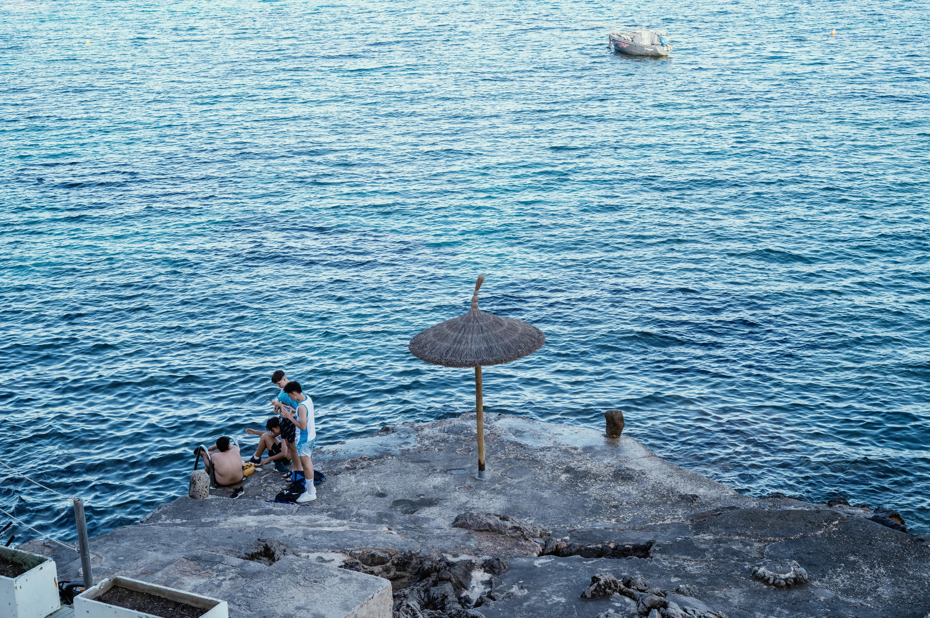 Group of people on a rocky shore next to a straw umbrella and calm blue sea.