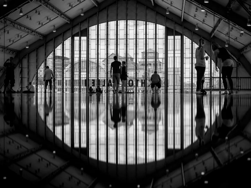 Silhouetted people in an indoor space with floor reflections and large windows showing a cityscape.