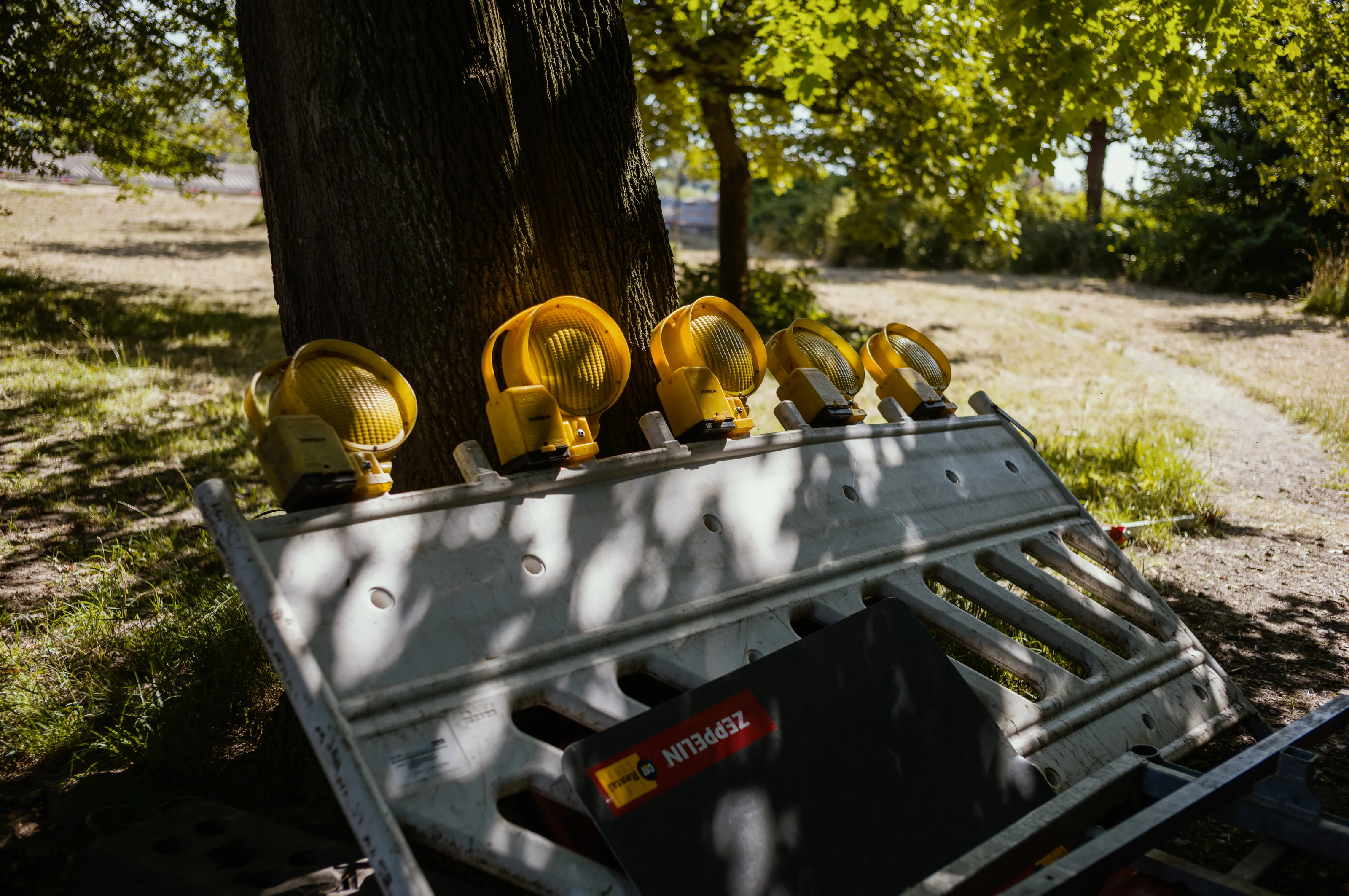 A row of yellow construction lights resting on a metal barricade under a tree.