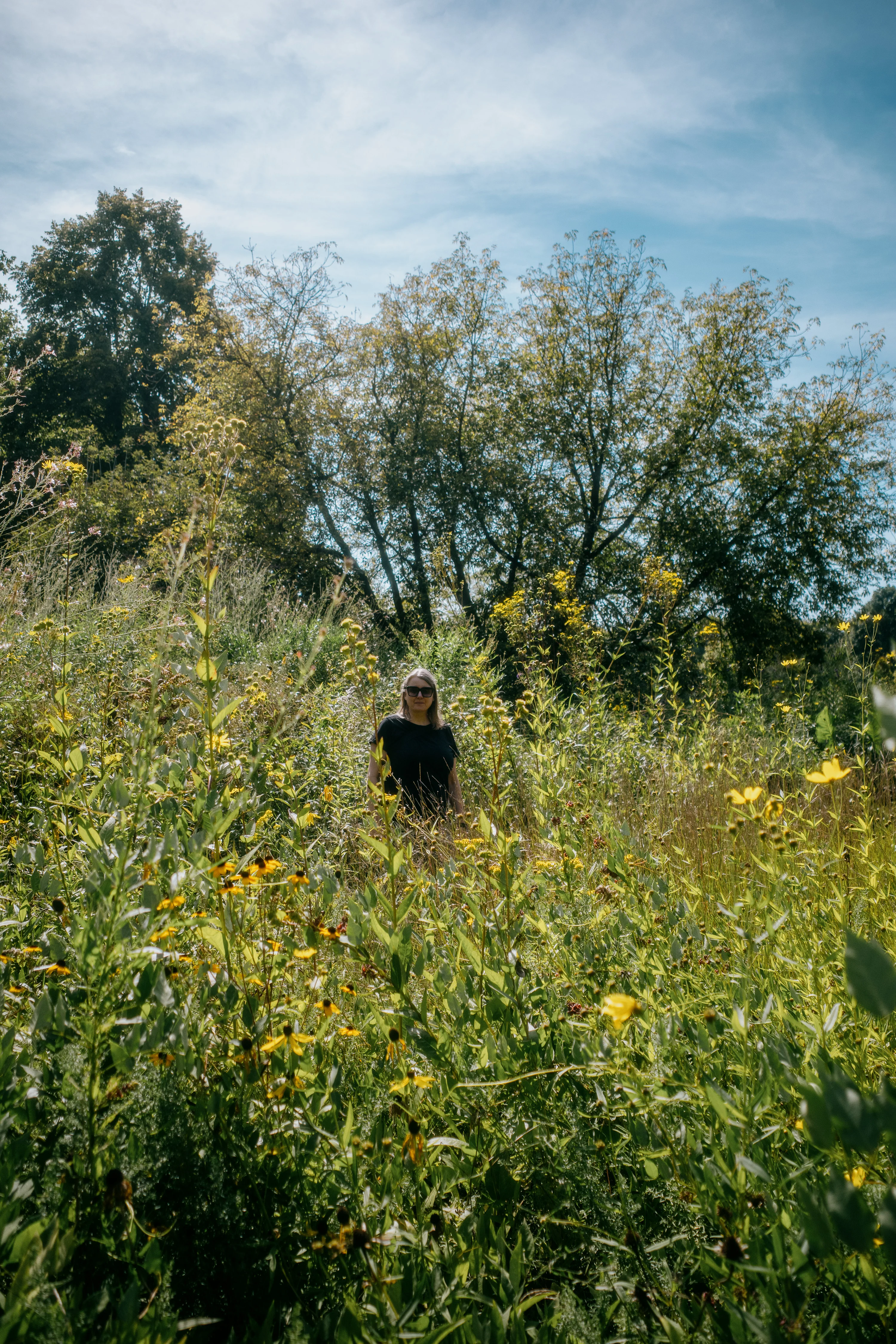 Person standing in a field of tall wildflowers under a blue sky.