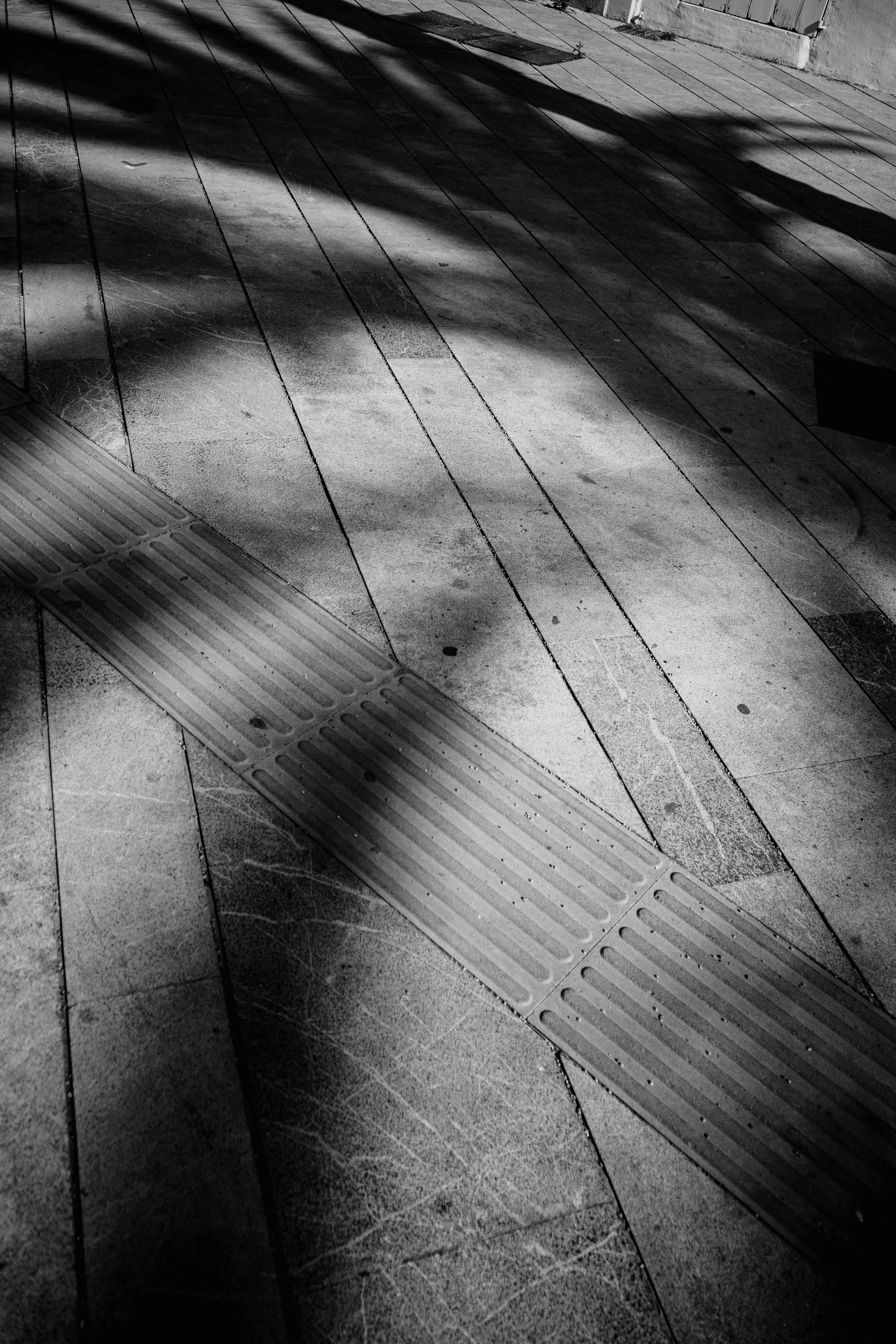A black and white image of a textured sidewalk with shadows and a tactile paving strip.