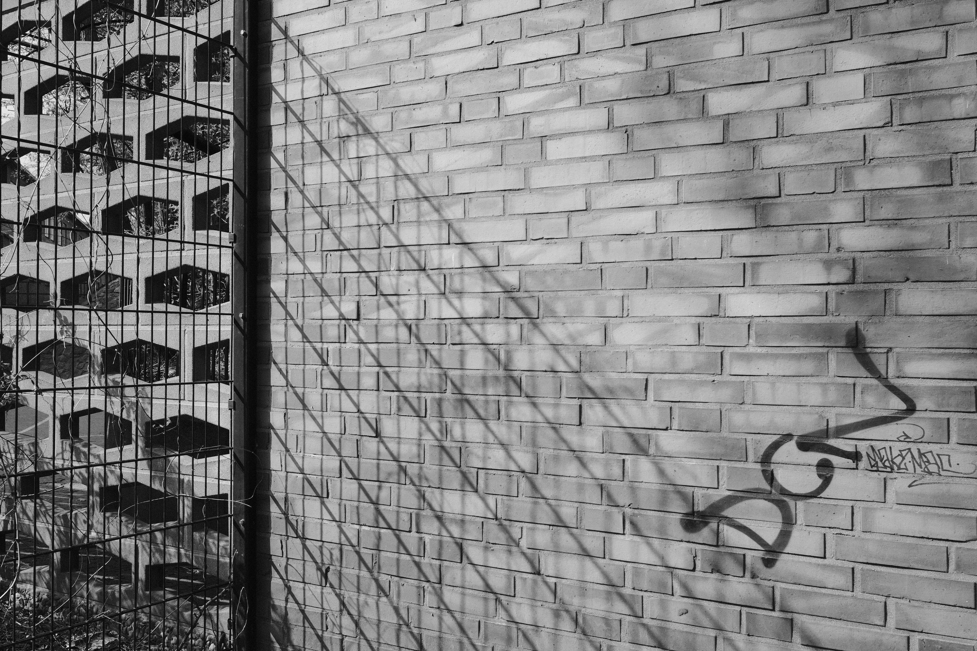 A brick wall with graffiti and the shadow of a metal fence cast across it.