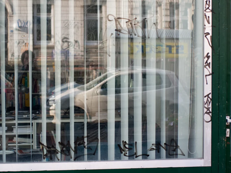 A car reflected in a glass window with vertical blinds and graffiti.