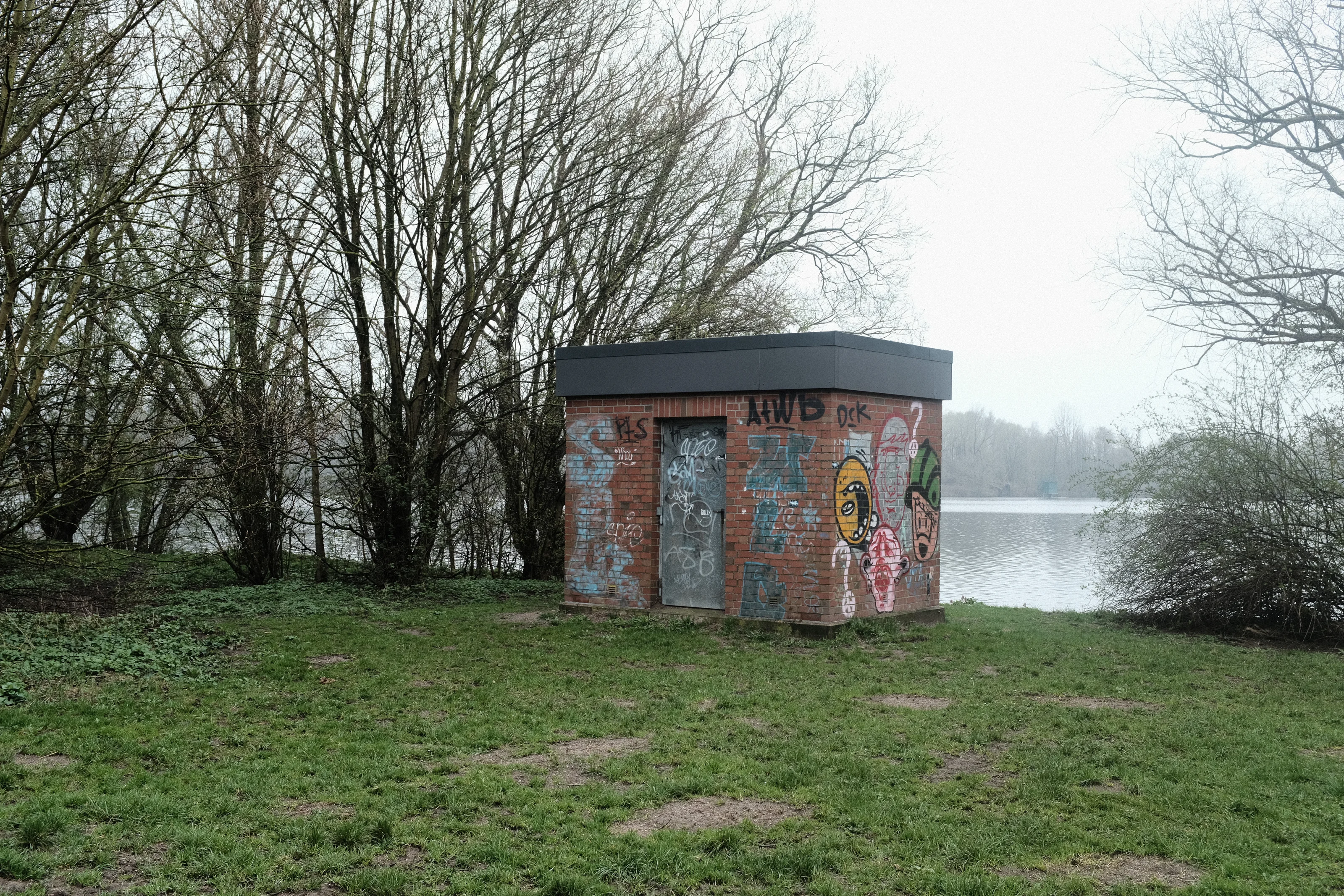Small graffiti-covered brick building beside a lake with surrounding trees.