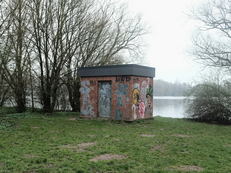 Small graffiti-covered brick building beside a lake with surrounding trees.