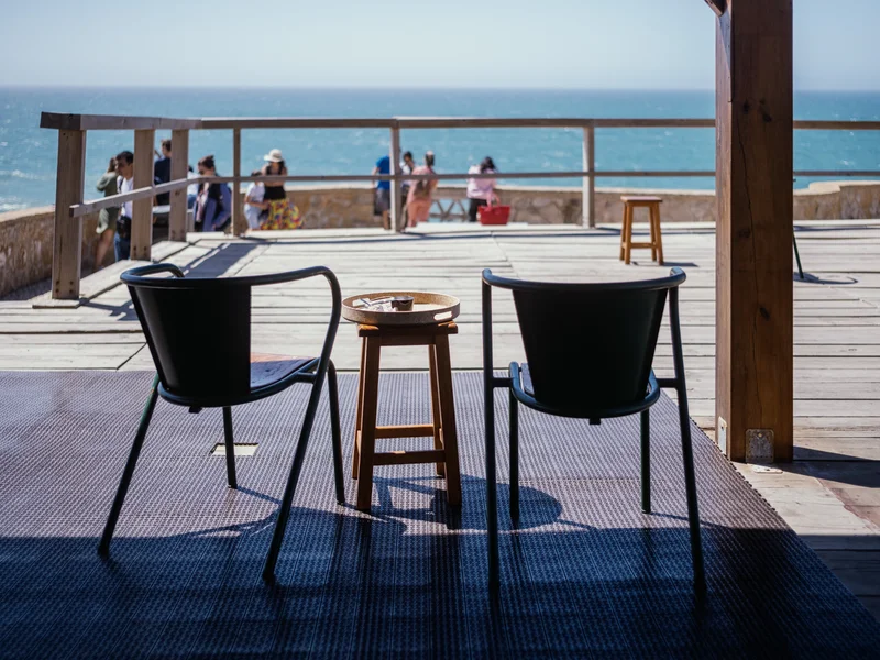 Two chairs and a small table facing the sea with people in the background on a deck.