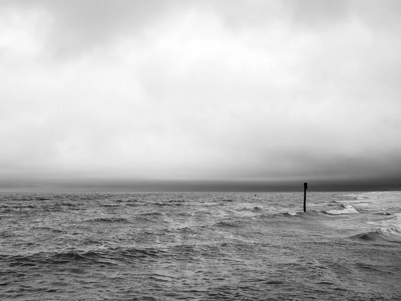 Black and white photo of a vast ocean with a solitary buoy in the distance under a cloudy sky.