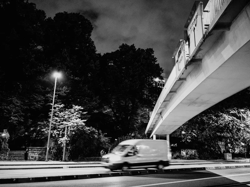 A van driving under a bridge at night with streetlights illuminating trees.