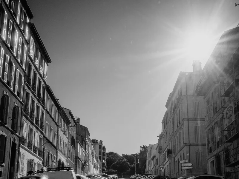 A sunlit street between rows of buildings with parked cars, casting long shadows.