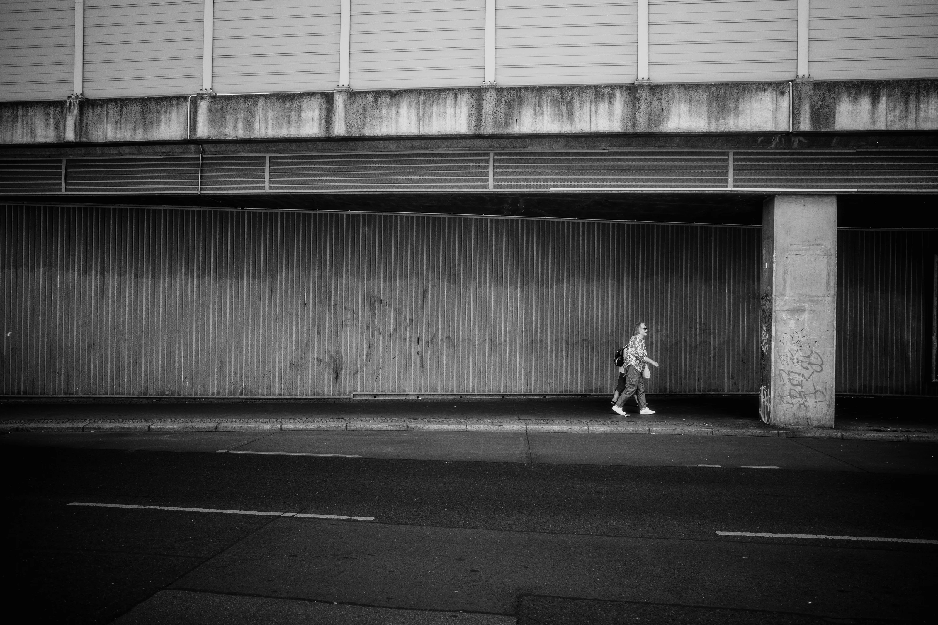 A person walking along a sidewalk under a large concrete structure.