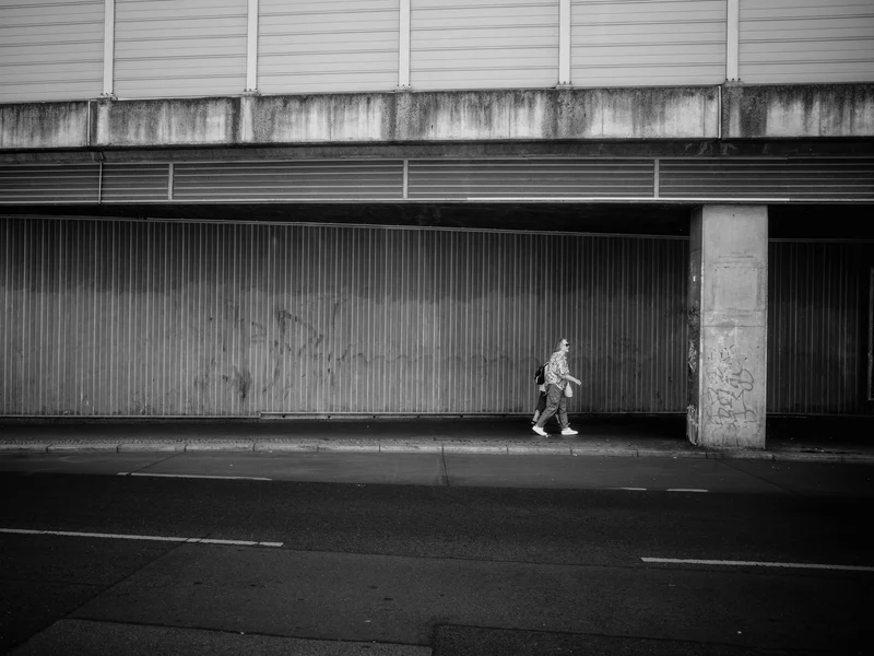 A person walking along a sidewalk under a large concrete structure.